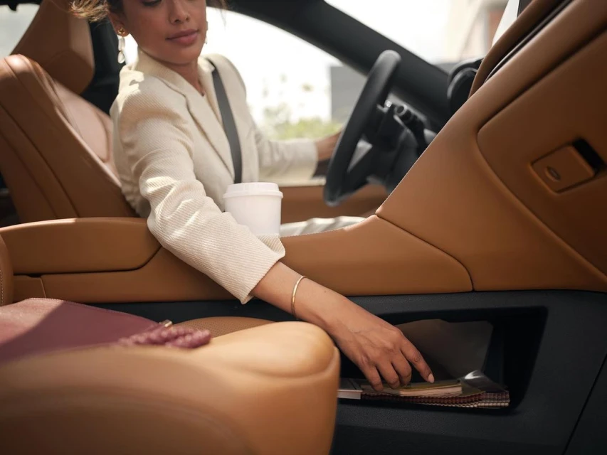 Professional woman driving car in business attire with coffee cup, reaching for items in the side compartment; focus on interior details.