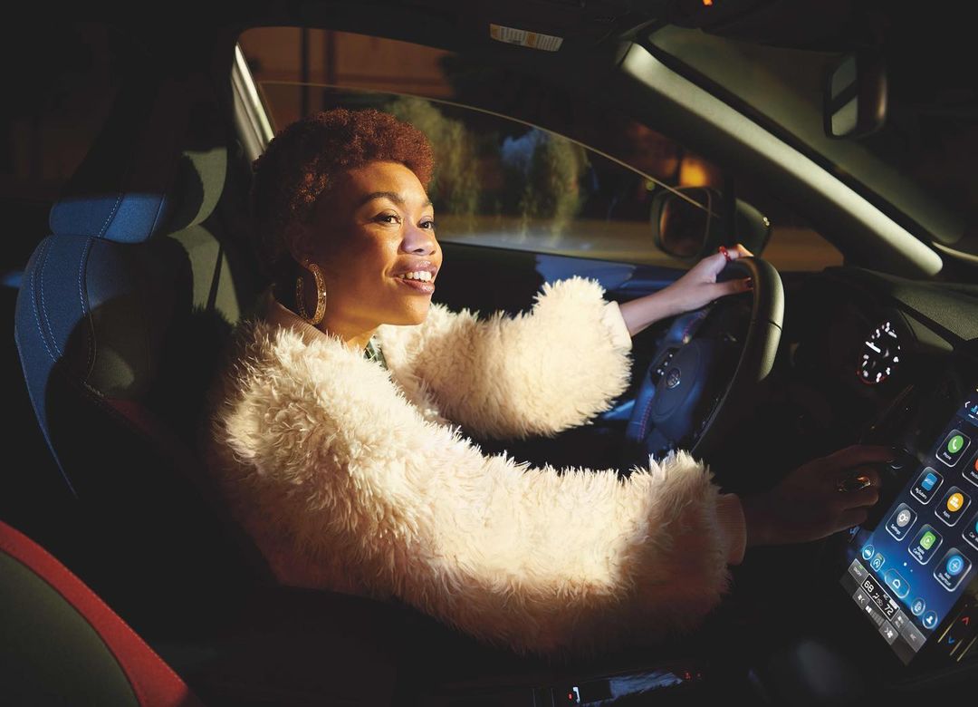 Smiling woman drives a car at night, illuminated by the car's interior lights, focusing on her cheerful expression and modern dashboard.
