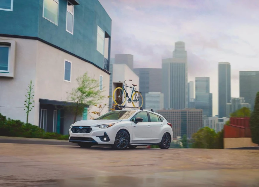 White Subaru Impreza with a bicycle mounted on the roof rack, parked on a street with city skyline in the background on a cloudy day.