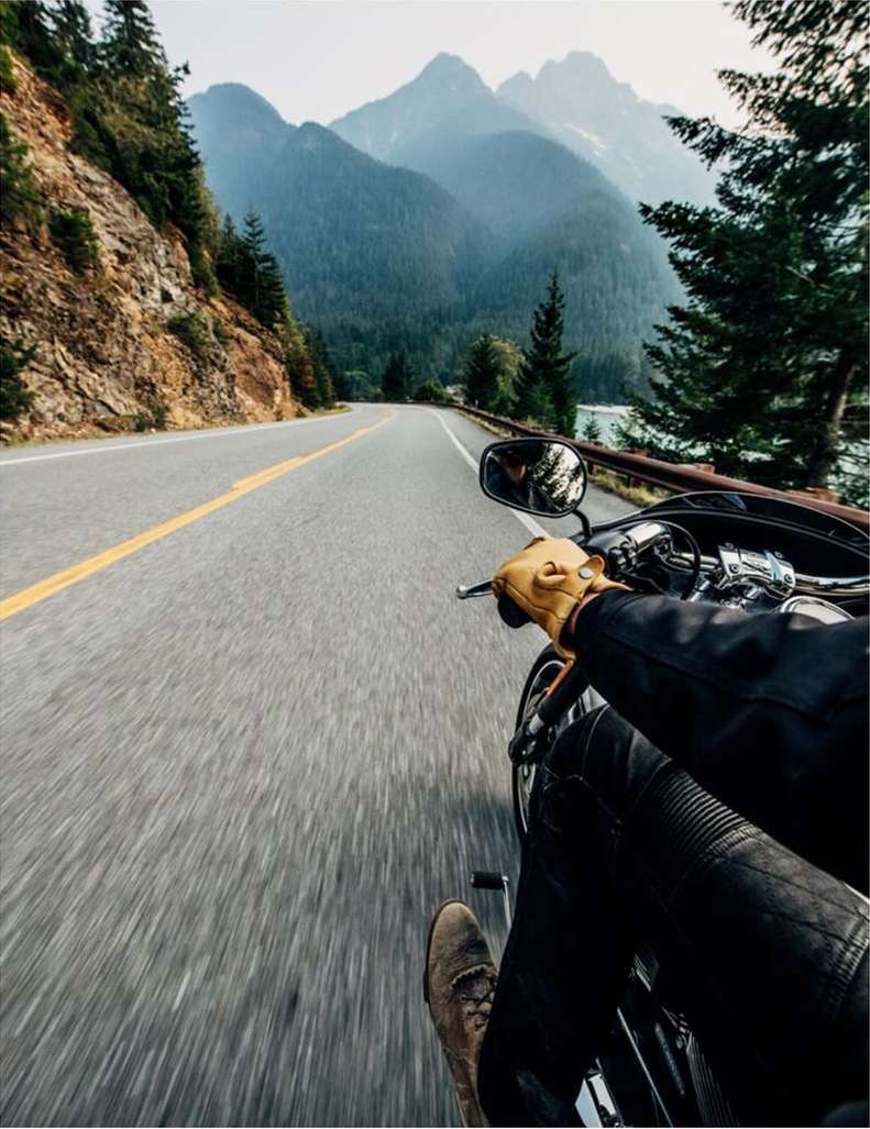 Motorcycle rider on open asphalt road flanked by tree-covered cliffside and hazy mountain range under overcast sky, first-person view.