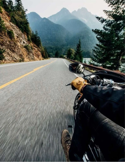 Motorcycle rider on open asphalt road flanked by tree-covered cliffside and hazy mountain range under overcast sky, first-person view.