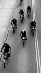 High-angle black and white photograph of a group of motorcyclists riding down an asphalt street lined with double white lines.
