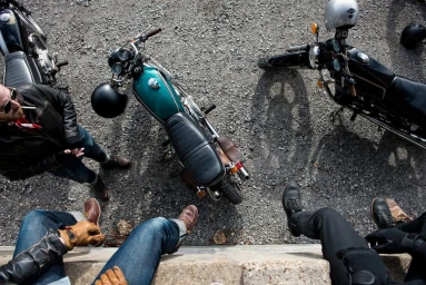 High-angle shot of three vintage motorcycles parked on gravel with riders sitting nearby.