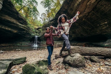 Man holding hands with a smiling woman balancing on a mossy fallen log near a waterfall in a rocky gorge, photographed by Yve Assad.