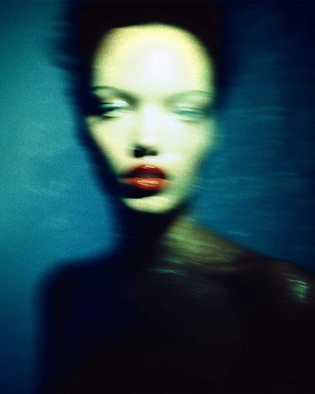 Abstract studio portrait of a woman with pale skin and bright red lips against a blue backdrop, captured with intentional motion blur.
