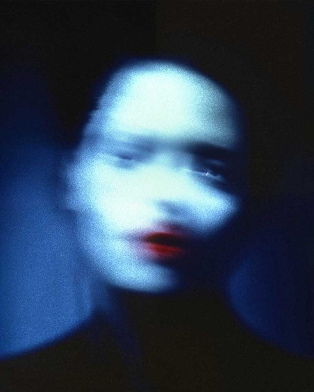 A blurred close-up of a female face with bright red lipstick, with a dreamy, ethereal quality in shades of blue and black tones.