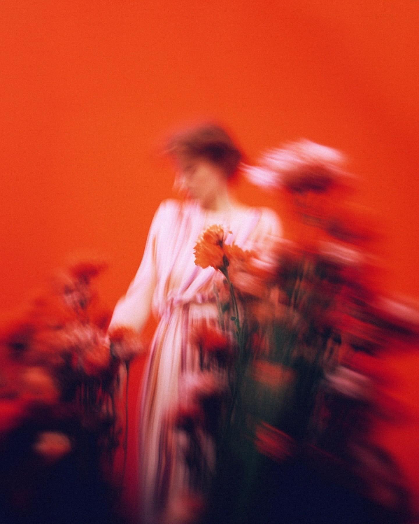 Blurred motion portrait of a woman in a dress holding orange flowers, set against an orange studio background with moody lighting.