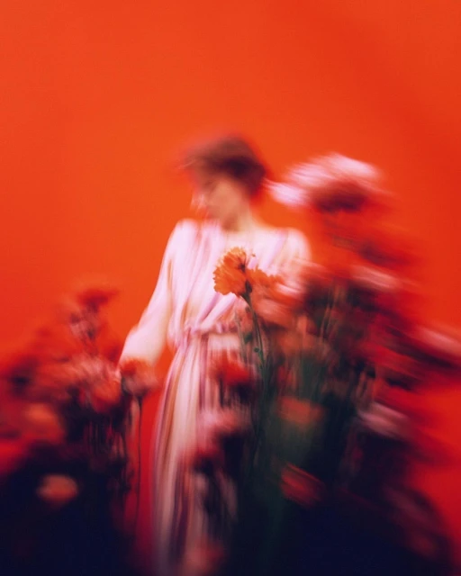 Blurred motion portrait of a woman in a dress holding orange flowers, set against an orange studio background with moody lighting.