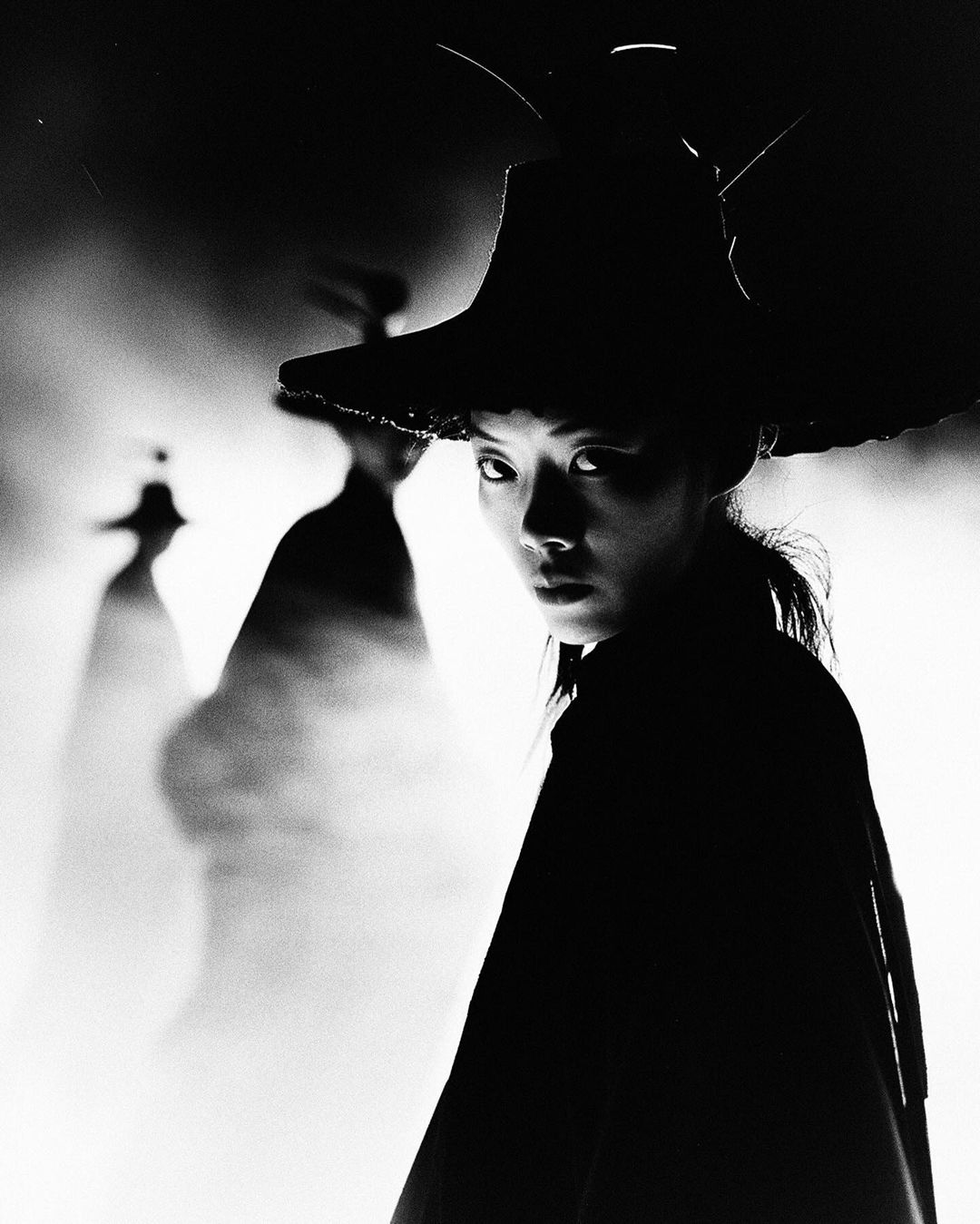 Striking black and white portrait of an Asian woman in a theatrical hat against a soft, smoky backdrop, creating a dramatic, artistic image.