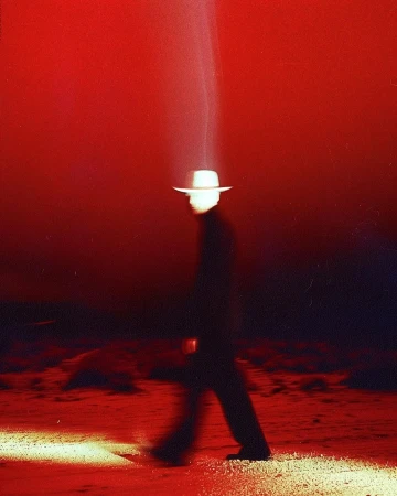 Blurred figure in a dark suit and white hat walking through a red-tinted, barren landscape under a spotlight effect.
