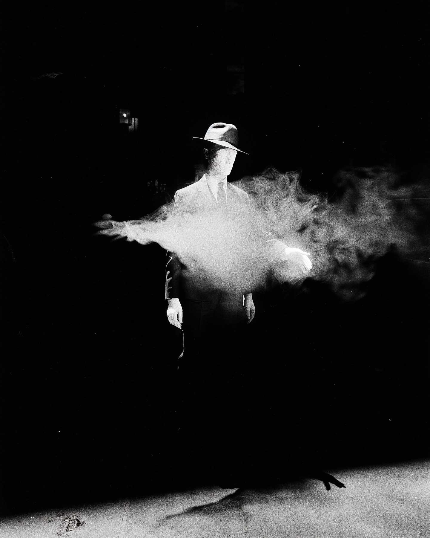 Monochrome portrait of a man in a suit and fedora standing in the dark with smoke around him, creating a mysterious atmosphere.