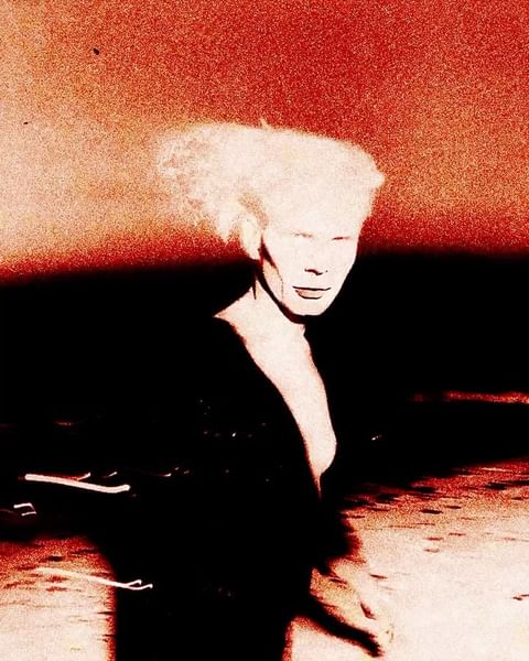 Woman with white hair in dark coat stands against an orange sky in a high-contrast artistic photograph with motion blur.