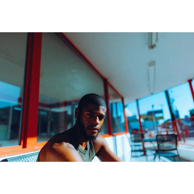 Adult male poses outdoors in a green tank top, seated near windows with a red frame, creating a casual, sunlit portrait.