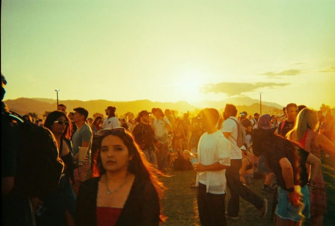Crowd of people gathered outdoors at sunset. Numerous adults are visible in a field as they enjoy a music festival.
