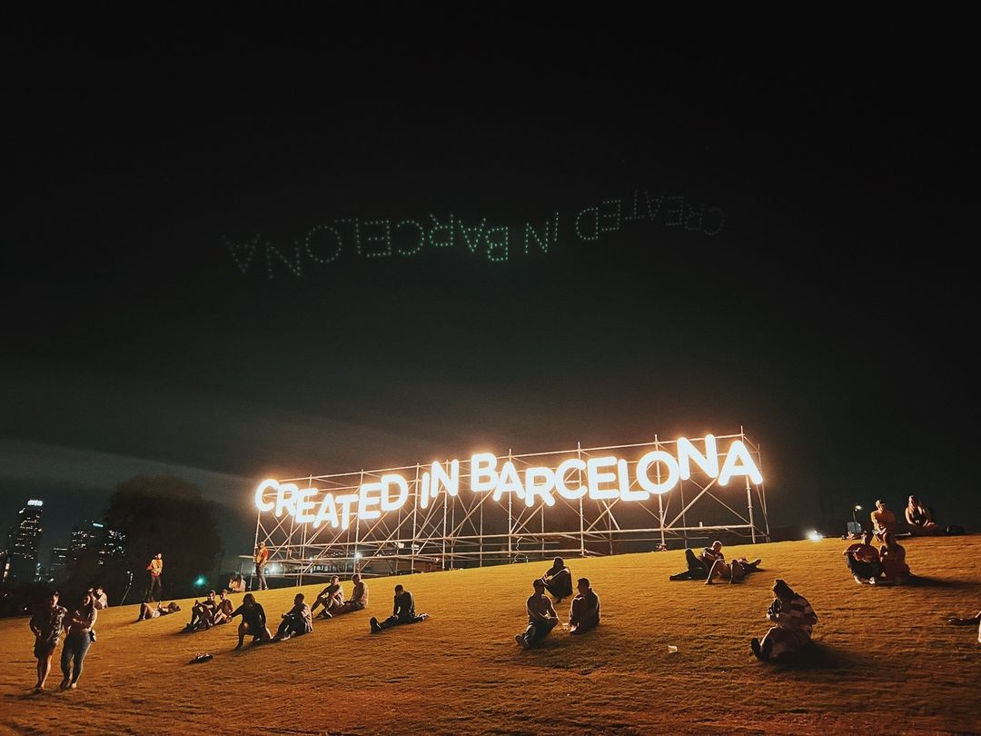 Large 'Created in Barcelona' sign glows above a grassy hill where a group of people relax at night with cityscape background.