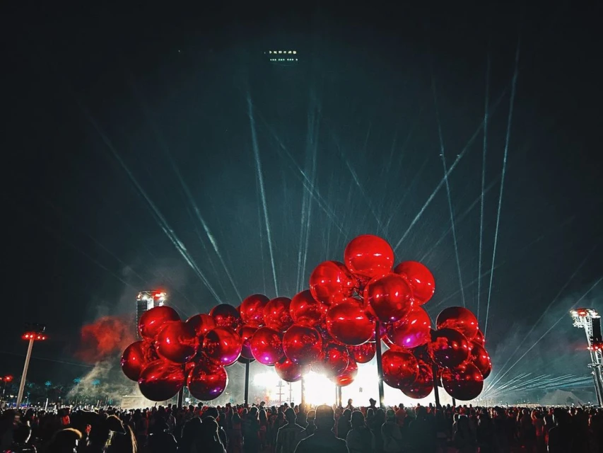 A large group of red balloons float above a crowd of people at an outdoor event at night with bright lights shining into the sky.