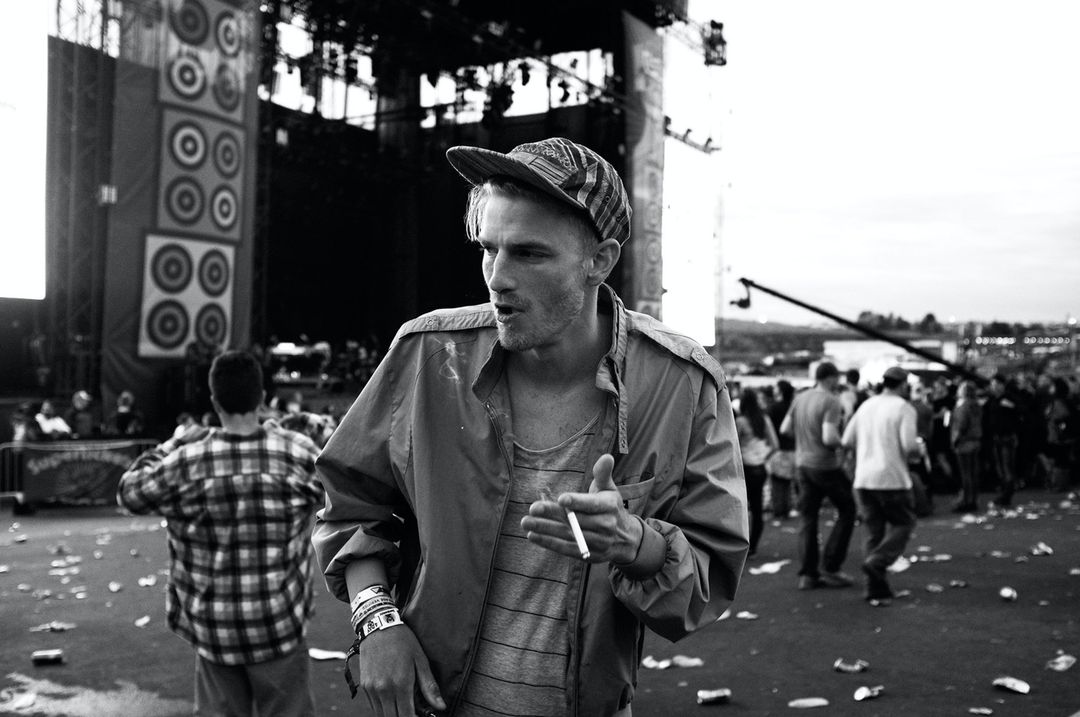 Black and white outdoor shot of a man with a baseball cap and jacket holding a cigarette, with a crowd and stage in the background.