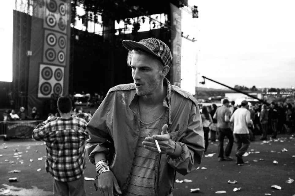 Black and white outdoor shot of a man with a baseball cap and jacket holding a cigarette, with a crowd and stage in the background.