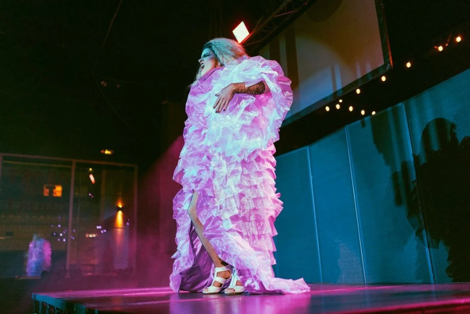 Drag queen in a pink ruffled gown stands on a stage with pink and blue lighting, showcasing an expressive performance and bold fashion.