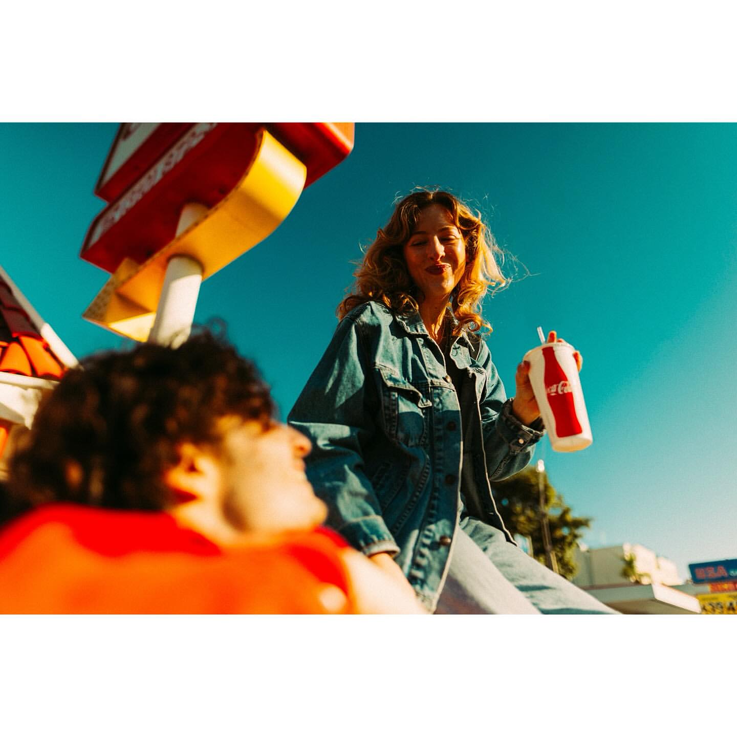 Two people outside a roadside diner with a classic Coca-Cola cup and vintage feel against a clear blue sky and building signage.