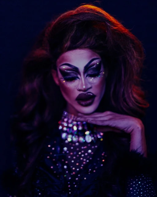 Glamour portrait of a drag queen with dramatic makeup and long auburn hair, wearing a jeweled dress, posing against a dark backdrop.