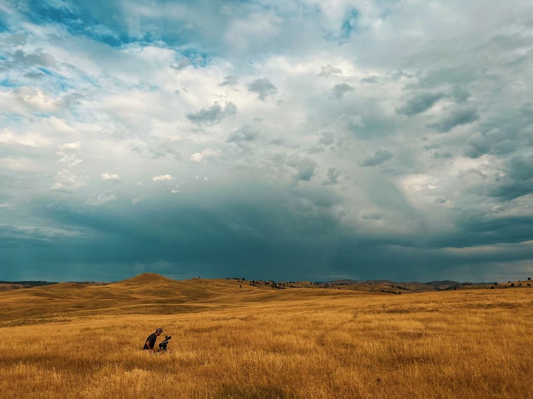 Photographer kneeling in a golden grassy field, capturing nature under a partly cloudy sky with hills in the background.