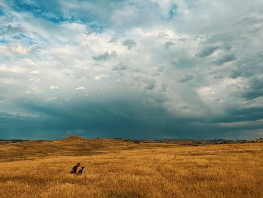 Photographer kneeling in a golden grassy field, capturing nature under a partly cloudy sky with hills in the background.