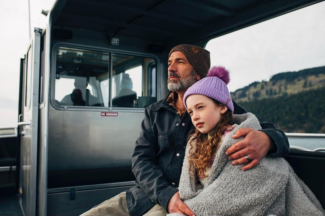 A man and a young girl are seated together on a boat, the girl wrapped in a gray blanket, with a scenic background of wooded hills.