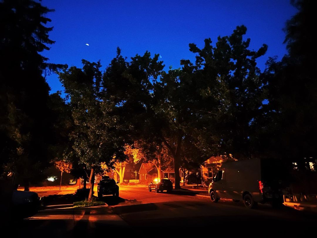 Night view of a suburban street with cars parked along the road, houses lit from within, and trees silhouetted against a blue sky.