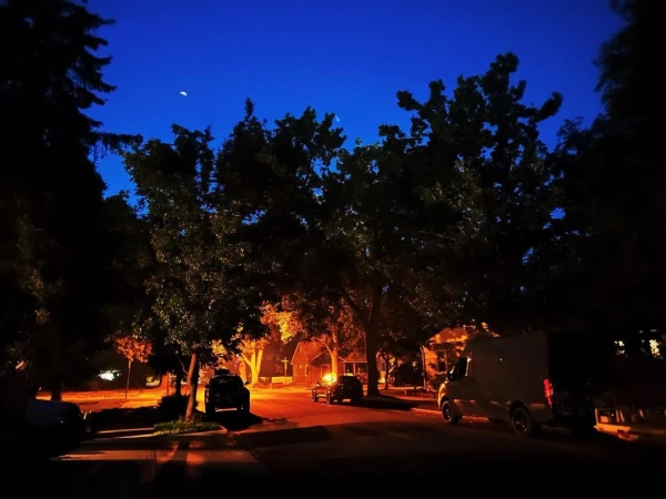 Night view of a suburban street with cars parked along the road, houses lit from within, and trees silhouetted against a blue sky.