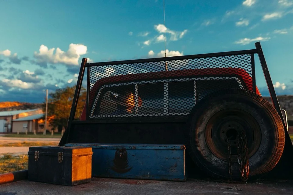 Truck bed with spare tire, tool boxes, and metal grate under a blue sky with clouds in a rural setting during golden hour.