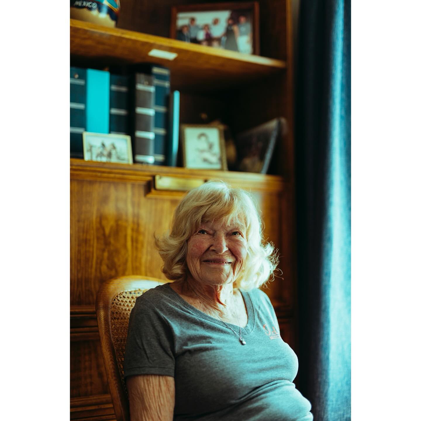 Portrait of a smiling elderly woman with short blonde hair sitting in a chair, with a wood bookshelf and blue curtains in background.