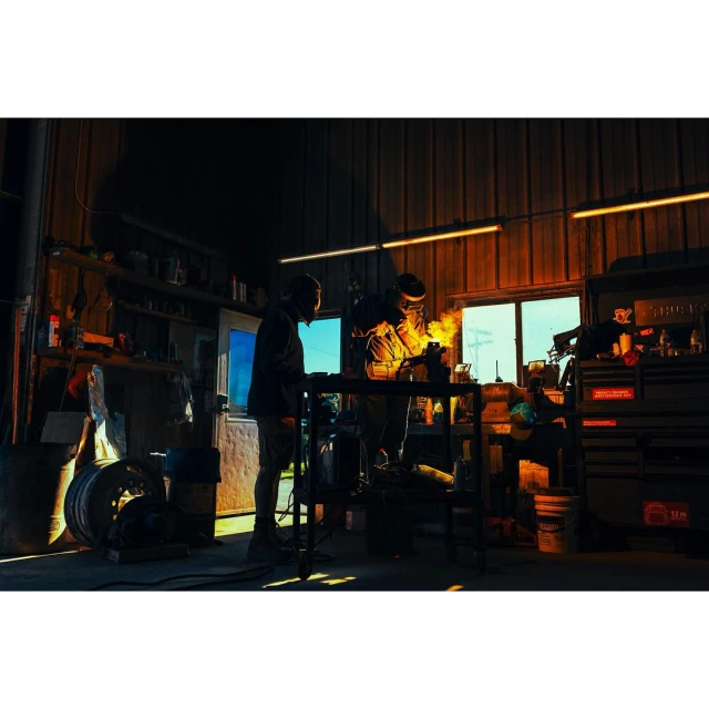 Two welders in a dimly lit garage work on a metal project, sparks flying from their tools under a warm-toned lighting.