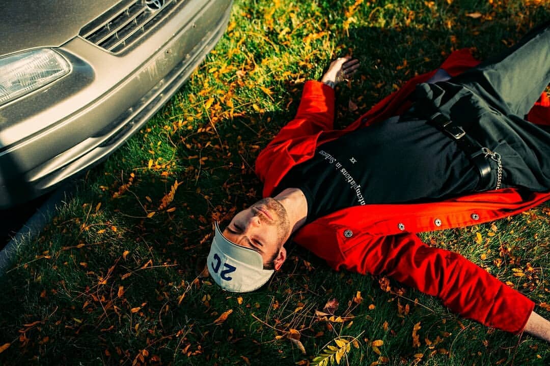 Adult male model in black t-shirt and red jacket lays on grass with fallen leaves near silver car in outdoor, sunny daytime scene.