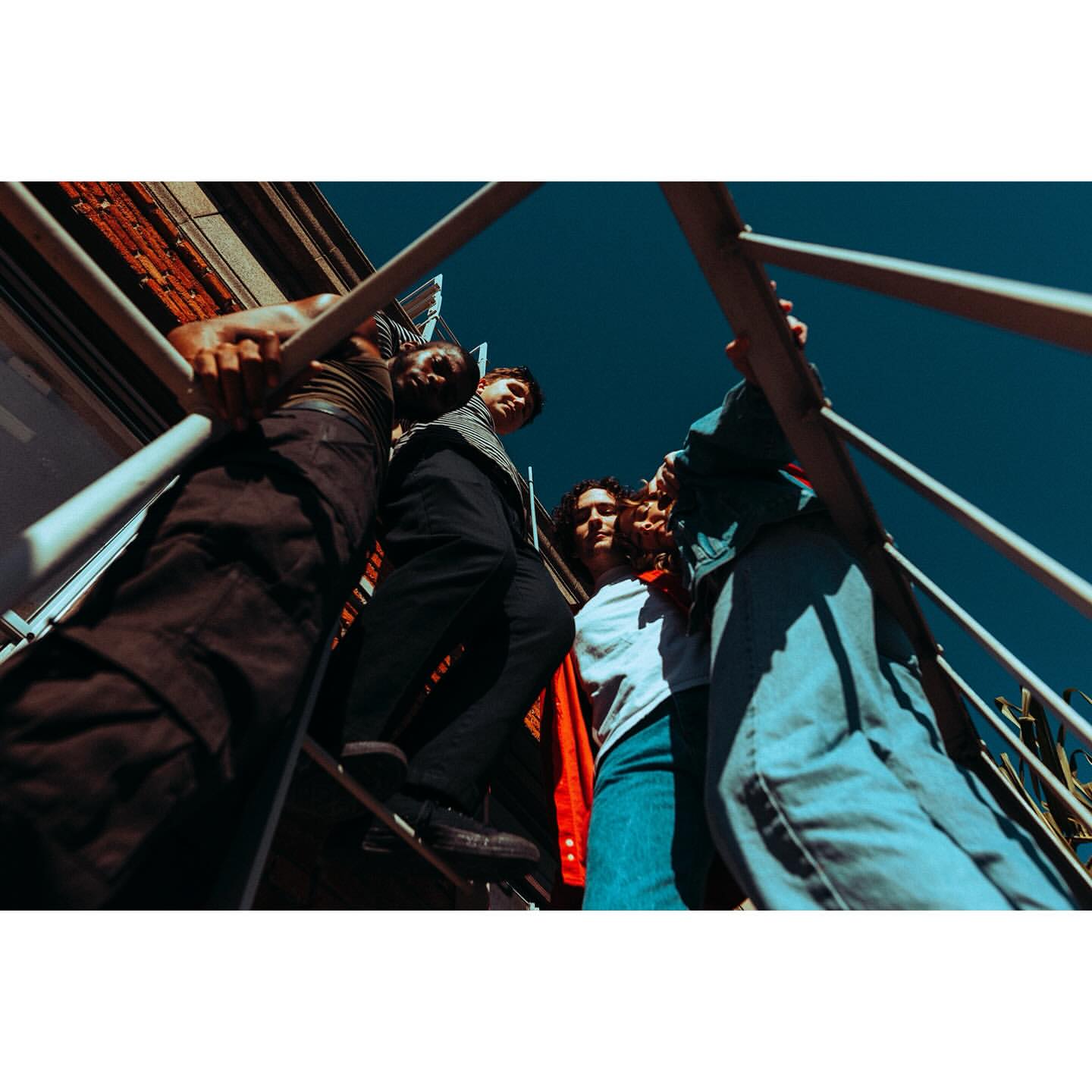 Low-angle view of a group of young adult friends on a metal staircase against a blue sky, creating a dynamic, urban fashion editorial.
