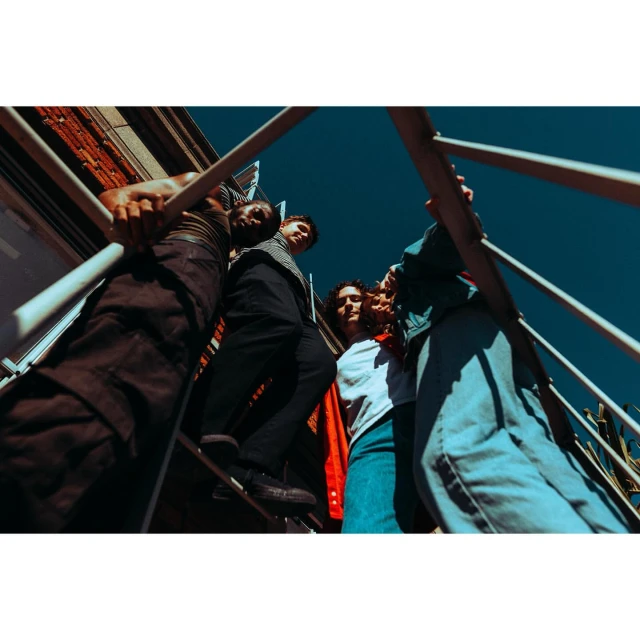 Low-angle view of a group of young adult friends on a metal staircase against a blue sky, creating a dynamic, urban fashion editorial.
