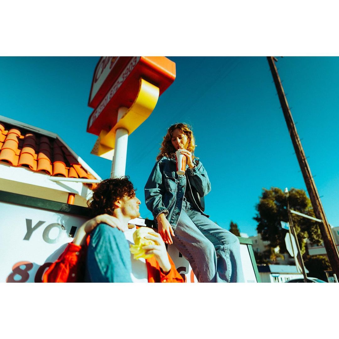 Two young adults in denim sit outdoors; one drinks from a cup, the other eats, with a fast-food restaurant and blue sky in the background.