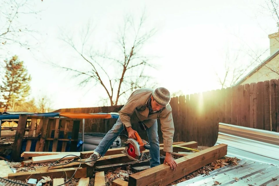 Man wearing a cap is cutting a wood plank outside with a power saw in daytime, with house roof, trees, and fence visible.
