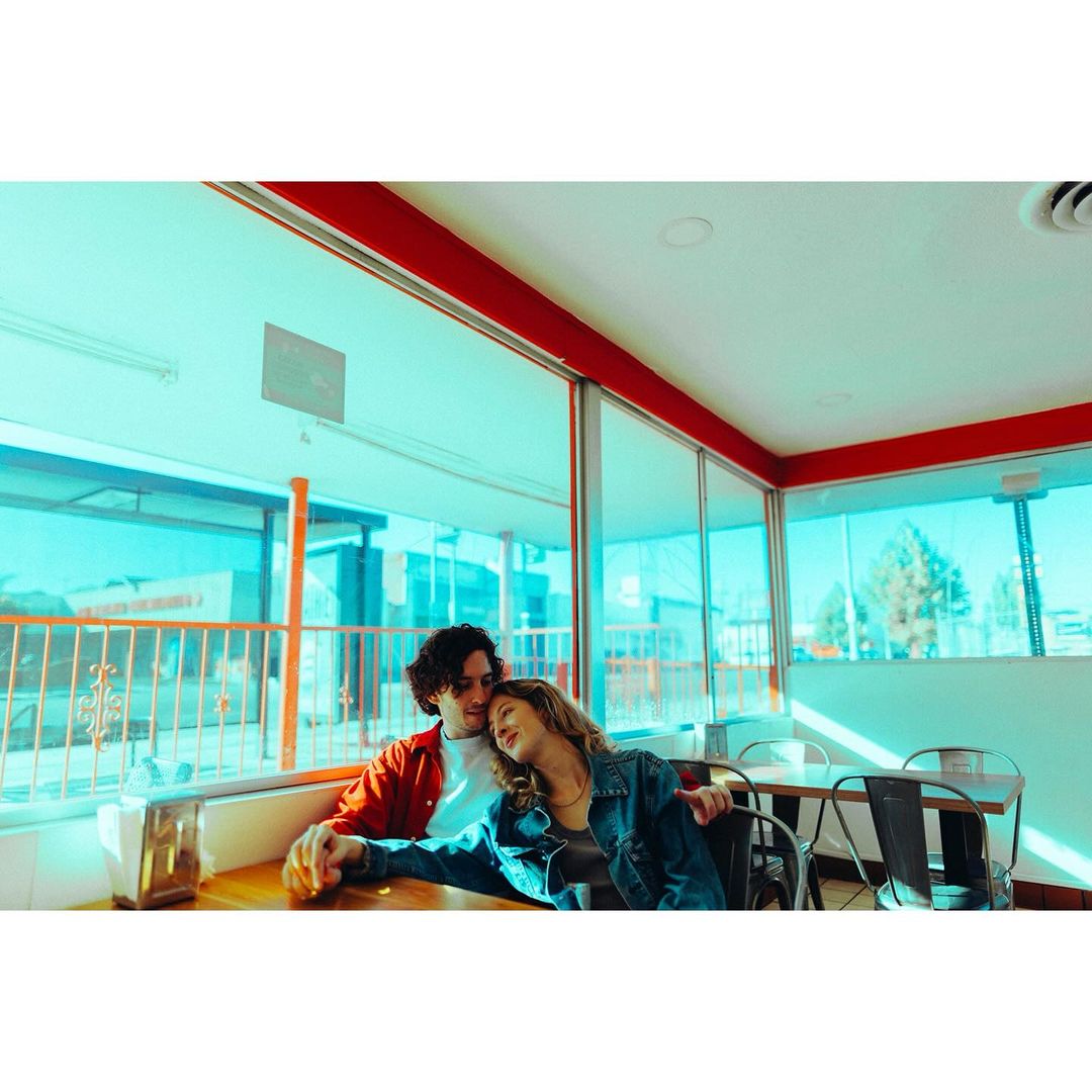 Couple in casual attire sitting at a table in a vintage diner with natural window light creating a cozy, intimate atmosphere. 