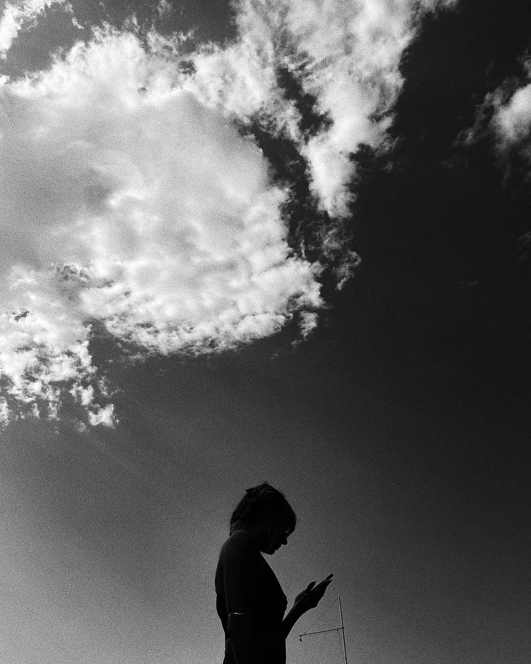 Silhouette of a person holding a smartphone against a sky with dramatic clouds in a black and white photograph.