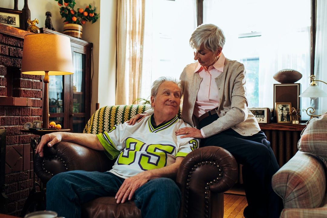 Elderly couple in living room: senior man sits in leather chair with woman's hand on his shoulder, family photos in background.