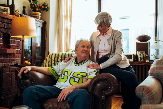 Elderly couple in living room: senior man sits in leather chair with woman's hand on his shoulder, family photos in background.