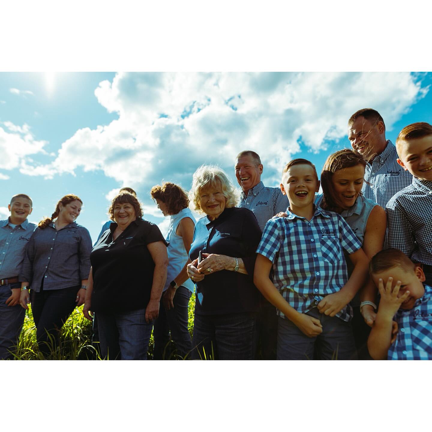 A group of people stands outside with blue sky and clouds behind them. There are children and adults in the photo. Everyone is smiling.