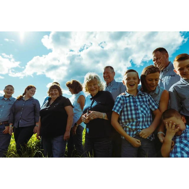 A group of people stands outside with blue sky and clouds behind them. There are children and adults in the photo. Everyone is smiling.