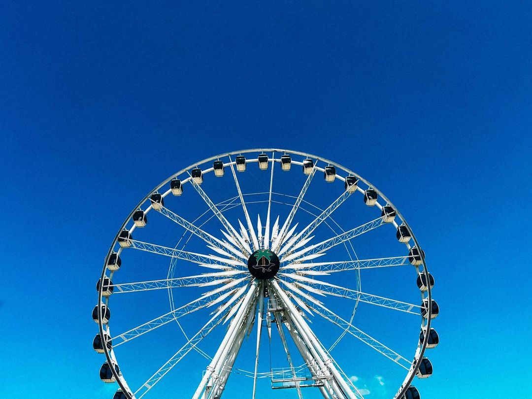 A white metal ferris wheel stands against a bright blue sky, showcasing its intricate structure and geometric design on a sunny day.