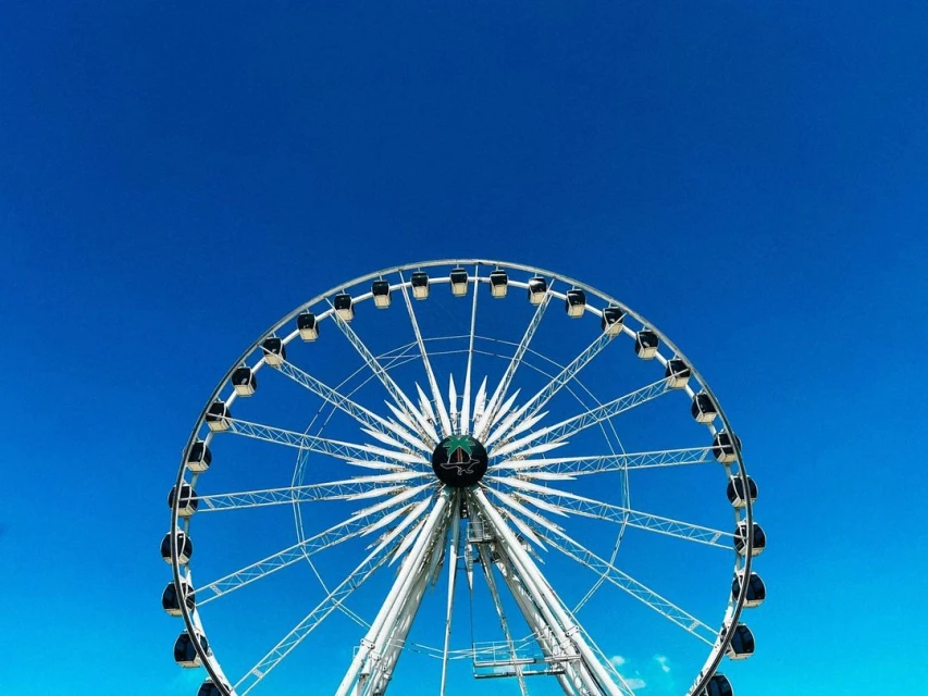 A white metal ferris wheel stands against a bright blue sky, showcasing its intricate structure and geometric design on a sunny day.