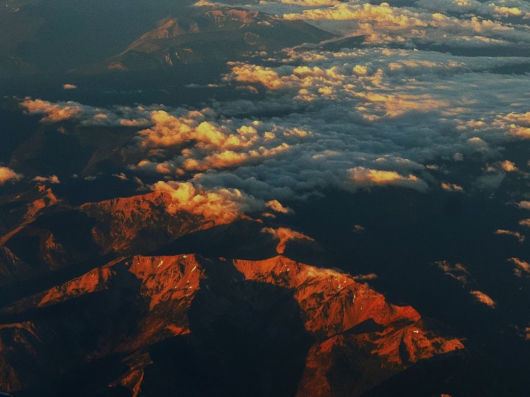Aerial view of rugged mountain range peaks with snow and rock textures, partially covered by a layer of clouds during golden hour.