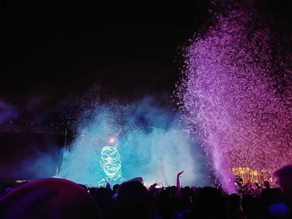 Crowd at an outdoor music concert with a large digital face projected on the stage with confetti and lights against a dark sky.