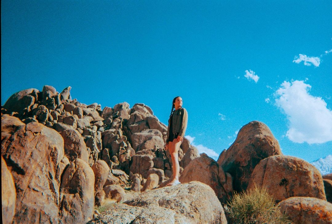 Young woman standing on large rocks in a natural desert landscape, against a clear blue sky with scattered clouds on a sunny day.