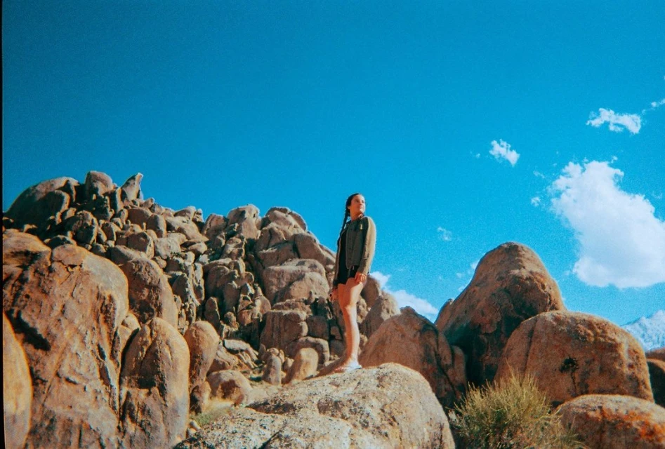 Young woman standing on large rocks in a natural desert landscape, against a clear blue sky with scattered clouds on a sunny day.